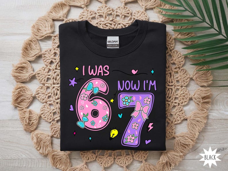 May include: A black t-shirt featuring a graphic design with the text I WAS NOW I'M and the numbers 6 and 7. The number 6 is pink with floral patterns and blue bows, while the number 7 is purple with floral patterns and a pink bow, surrounded by small icons.