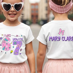 May include: A white short-sleeved t-shirt featuring a graphic print. The front displays the text I WAS 6 NOW I'M 7 with pink and purple numbers decorated with flowers and bows. The back features the name MARY CLARE in purple with a pink bow.