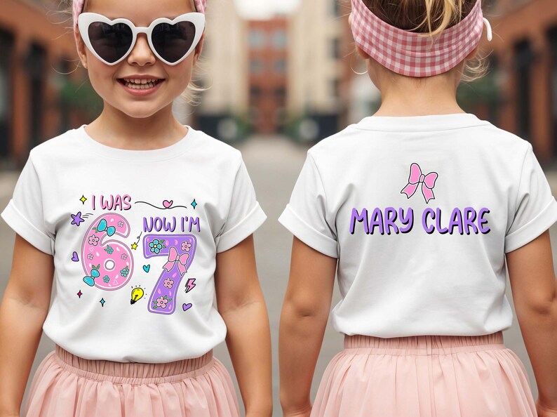 May include: A white short-sleeved t-shirt featuring a graphic print. The front displays the text I WAS 6 NOW I'M 7 with pink and purple numbers decorated with flowers and bows. The back features the name MARY CLARE in purple with a pink bow.