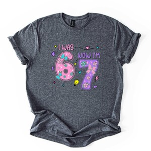 May include: A dark heather grey short-sleeve t-shirt featuring the text I WAS NOW I'M and the numbers 6 and 7. The number 6 is pink with blue bows and flowers, and the number 7 is purple with a pink bow and flowers, surrounded by small icons.
