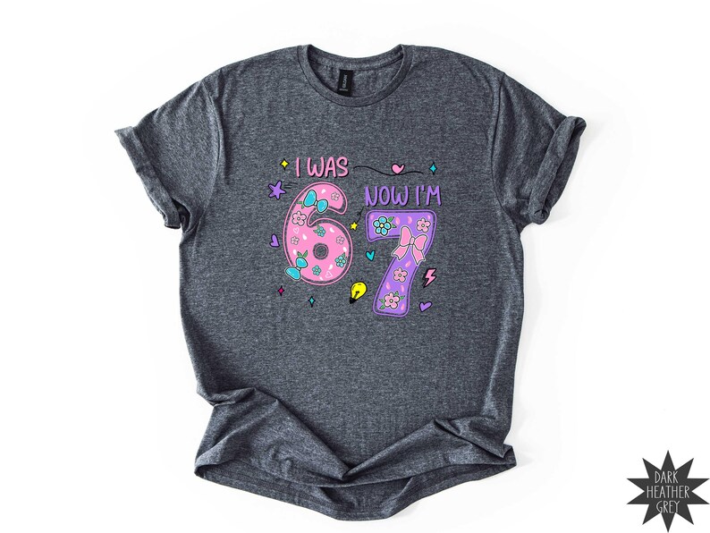 May include: A dark heather grey short-sleeve t-shirt featuring the text I WAS NOW I'M and the numbers 6 and 7. The number 6 is pink with blue bows and flowers, and the number 7 is purple with a pink bow and flowers, surrounded by small icons.