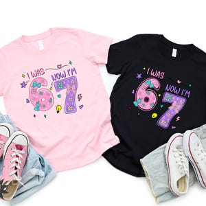 May include: A light pink t-shirt and a black t-shirt, each featuring a graphic with the text 'I WAS 6 NOW I'M 7' in a colorful, playful font. The design includes illustrations of bows, flowers, and stars. Each shirt is paired with blue denim shorts and sneakers.