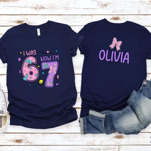 May include: Two navy blue t-shirts. The left shirt features the text "I WAS" and "NOW I'M" with a large pink "6" and purple "7" decorated with bows and flowers. The right shirt displays a pink bow and the name "OLIVIA" in purple lettering.