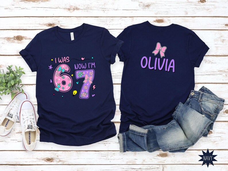 May include: Two navy blue t-shirts. The left shirt features the text "I WAS" and "NOW I'M" with a large pink "6" and purple "7" decorated with bows and flowers. The right shirt displays a pink bow and the name "OLIVIA" in purple lettering.