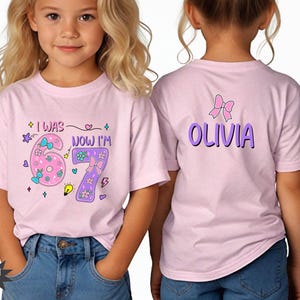 May include: A light pink t-shirt featuring a graphic on the front that reads I WAS 6 NOW I'M 7 with colorful flowers, bows, and stars. The back of the shirt displays the name OLIVIA in purple letters with a pink bow graphic positioned above the name.