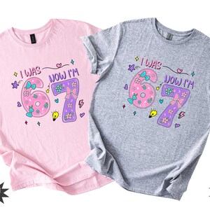 May include: A light pink t-shirt and a sport grey t-shirt, each featuring a graphic print with the text "I WAS" and "NOW I'M" surrounding the numbers 6 and 7, decorated with colorful bows, flowers, stars, a lightbulb, and lightning bolt icons.