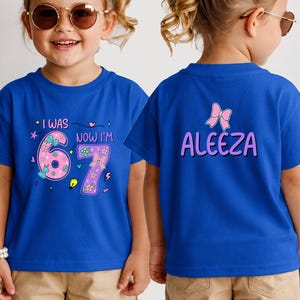 May include: A royal blue t-shirt featuring a front graphic with the text I WAS NOW I'M 6 7 and a back graphic with the name ALEEZA in purple letters. The front design includes pink numbers decorated with small flowers, bows, and colorful icons.