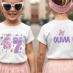 May include: A white short-sleeved t-shirt featuring a graphic print on the front and back. The front displays the text I WAS 6 NOW I'M 7 with pink and purple numbers decorated with bows and flowers. The back features the name OLIVIA in purple.