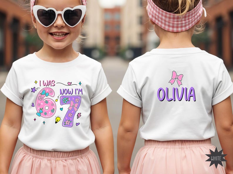 May include: A white short-sleeved t-shirt featuring a graphic print on the front and back. The front displays the text I WAS 6 NOW I'M 7 with pink and purple numbers decorated with bows and flowers. The back features the name OLIVIA in purple.