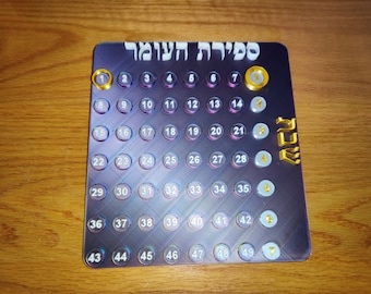 May include: A purple and silver counting board with the Hebrew text "Counting of the Omer" at the top. The board has 49 numbered circles, with gold accents on the numbers 1, 7, 14, 21, 28, 35, 42, and 49. The board is on a wooden surface.