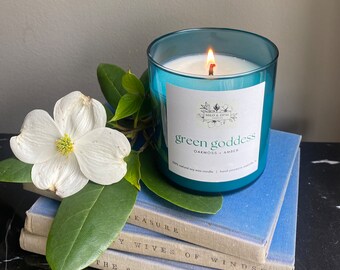 May include: A teal blue candle jar with a white label that reads "green goddess oakmoss + amber". The candle is lit and sitting on a stack of blue books.