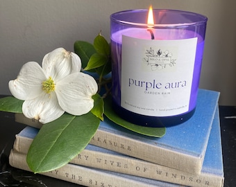 May include: Purple soy wax candle in a clear glass jar with a purple label that reads "Purple aura GARDEN RAIN 100% natural soy wax candle | hand poured in nashville, tn". The candle is sitting on a stack of books.