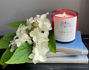 May include: A red glass candle jar with a white label that reads "ruby red grapefruit + mangosteen" and "MILO & OPAL SCENTS". The candle is sitting on a stack of books with the text "SURE", "ERRY WIVES OF", and "THE SIXTH: PART I" visible. White flowers are in the background.
