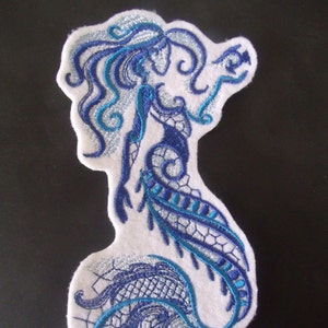 May include: Blue and white embroidered mermaid patch with intricate details. The mermaid is depicted with flowing hair and a tail with a lace-like design.