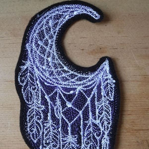 May include: A purple and white embroidered patch featuring a crescent moon with a dreamcatcher design. The patch has a white outline and white feathers hanging from the bottom.