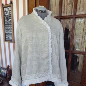May include: A white jacket with a green paisley pattern and white ruffle trim. The jacket has a shawl collar and long sleeves.