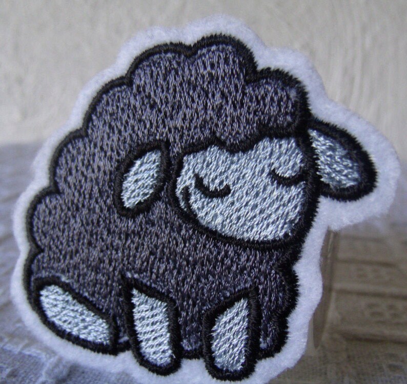 May include: A black embroidered sheep with a white background. The sheep has a closed-eye expression and is sitting on its hind legs.