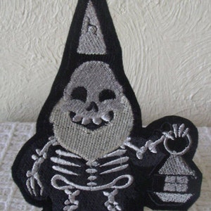May include: A black and white embroidered patch of a skeleton gnome wearing a pointed hat and holding a lantern. The gnome is smiling and has a skull for a head.