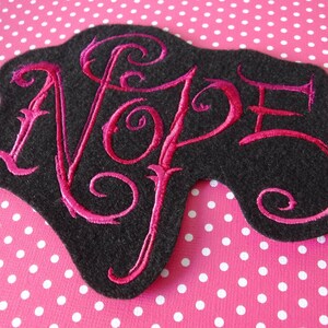 May include: A black felt patch with a pink embroidered word "NOPE" in a stylized font.