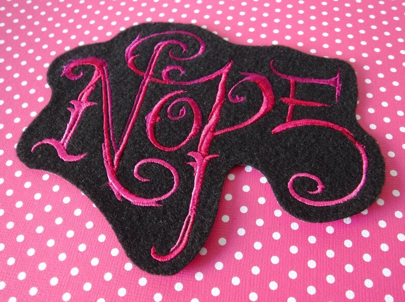 May include: A black felt patch with a pink embroidered word "NOPE" in a stylized font.