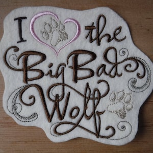 May include: Embroidered patch featuring the text "I love the Big Bad Wolf" in brown cursive lettering. A pink heart with a silver paw print is above the text. The patch is on a white background.
