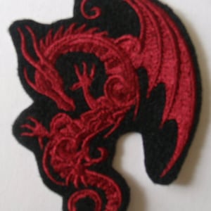 May include: Embroidered patch featuring a red dragon design on a black background. The dragon is depicted with intricate details, including wings and a serpentine body. The patch has a unique, irregular shape, suitable for clothing or accessories.