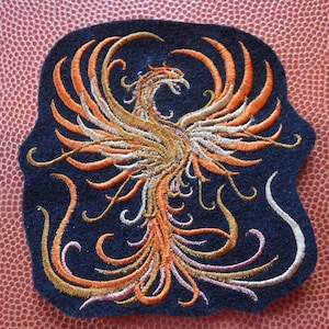 Peut inclure: Patch brodé représentant un phénix avec des plumes orange, dorées et blanches sur un fond bleu foncé. Le phénix est représenté dans un design stylisé et complexe.