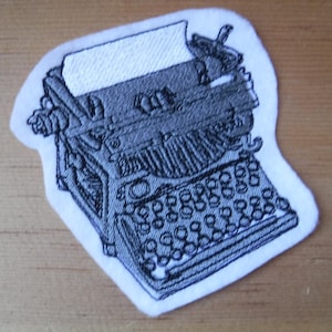 May include: A white fabric patch with an embroidered image of a vintage typewriter in black thread.