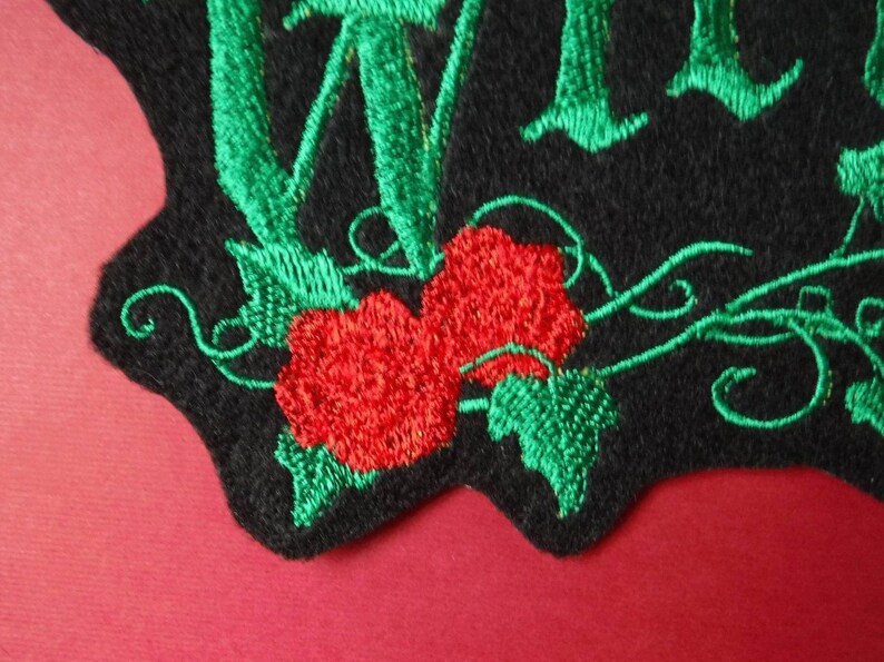 K&ouml;nnte beinhalten: Gestickter Aufn&auml;her mit gr&uuml;nen Bl&auml;ttern und roten Rosen auf schwarzem Hintergrund. Der Text "Wild" ist in Gr&uuml;n gestickt.