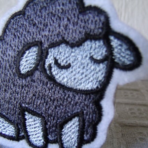 May include: A gray embroidered patch of a sheep with a white outline. The sheep has a closed-eye expression.