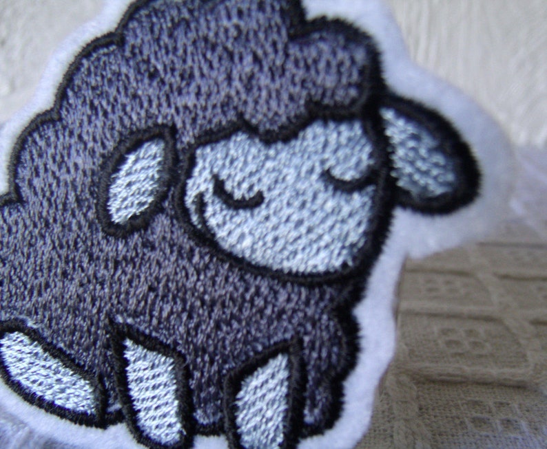 May include: A gray embroidered patch of a sheep with a white outline. The sheep has a closed-eye expression.