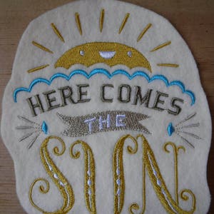 May include: A white felt patch with embroidered text that reads "Here comes the sun". The sun is yellow with a smiling face and blue rays. The text is in a decorative font with gold and silver thread.