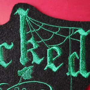 K&ouml;nnte beinhalten: Gr&uuml;ner gestickter Aufn&auml;her mit dem Wort "Wicked" in gotischer Schrift, mit einem Spinnennetz-Design im Hintergrund.