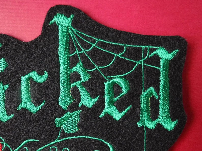 K&ouml;nnte beinhalten: Gr&uuml;ner gestickter Aufn&auml;her mit dem Wort "Wicked" in gotischer Schrift, mit einem Spinnennetz-Design im Hintergrund.