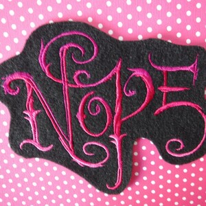 May include: A black felt patch with the word "NOPE" embroidered in pink cursive lettering.