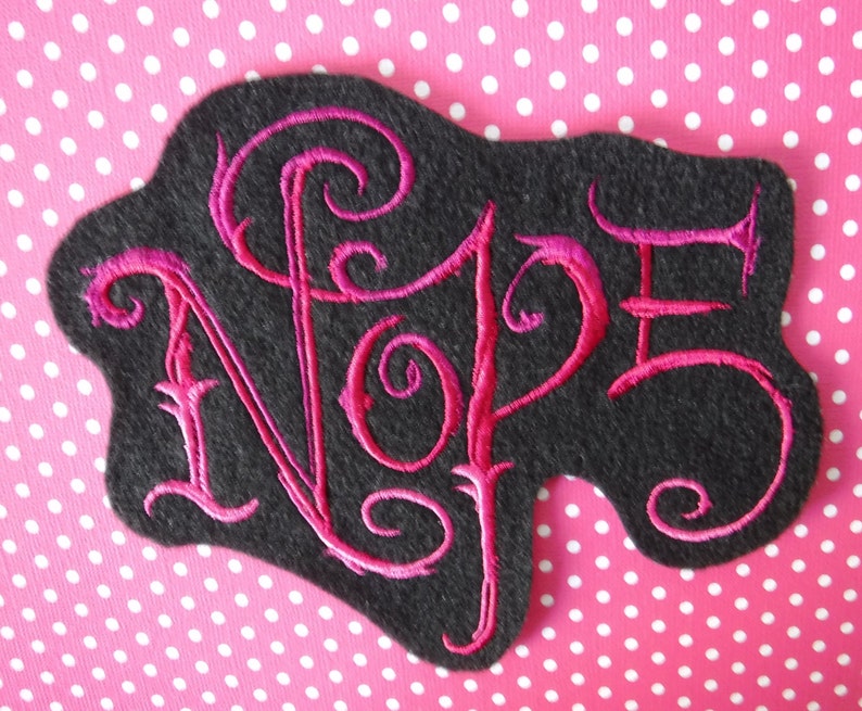 May include: A black felt patch with the word "NOPE" embroidered in pink cursive lettering.