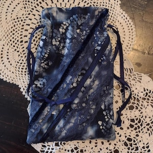 May include: A navy blue drawstring bag with a tie-dye pattern and lace accents. The bag features a dark blue ribbon drawstring closure and is set against a white crocheted doily.