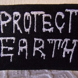 May include: Black rectangular patch with white embroidered text that reads "PROTECT EARTH". The letters have a dripping effect. The patch is made of a textured fabric.