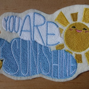 May include: Embroidered patch with a blue cloud and a yellow sun with a smiling face. The text "You are my sunshine" is embroidered in white on the patch.