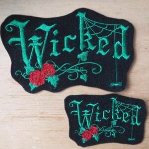 K&ouml;nnte beinhalten: Zwei schwarze Filzpatches mit gr&uuml;ner Stickerei. Der gr&ouml;&szlig;ere Patch zeigt das Wort "Wicked" in gotischer Schrift mit einem Spinnennetz und einer roten Rose. Der kleinere Patch hat das gleiche Design, aber ohne das Spinnennetz.