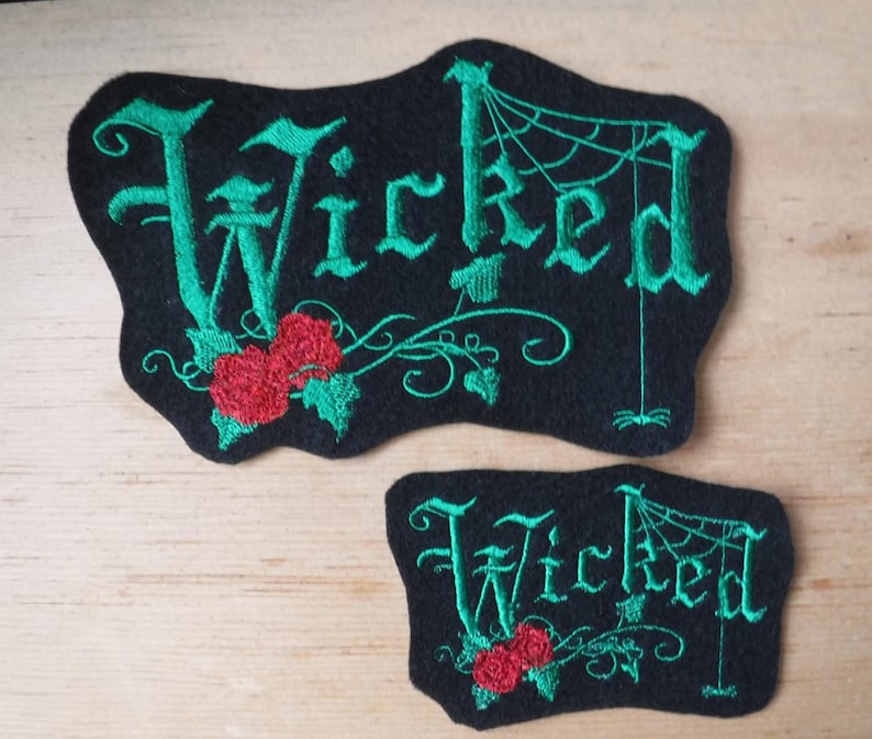K&ouml;nnte beinhalten: Zwei schwarze Filzpatches mit gr&uuml;ner Stickerei. Der gr&ouml;&szlig;ere Patch zeigt das Wort "Wicked" in gotischer Schrift mit einem Spinnennetz und einer roten Rose. Der kleinere Patch hat das gleiche Design, aber ohne das Spinnennetz.