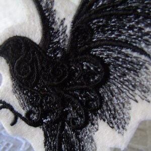 May include: Black embroidered raven design on a white fabric background. The raven is depicted with its wings spread out and its head turned to the side.