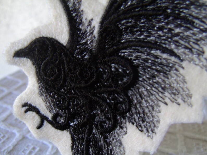 May include: Black embroidered raven design on a white fabric background. The raven is depicted with its wings spread out and its head turned to the side.