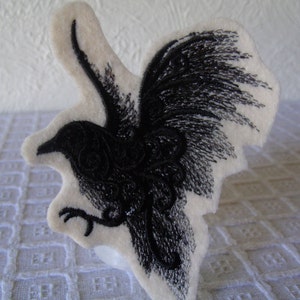 May include: Black embroidered crow with detailed wings on a white felt background.
