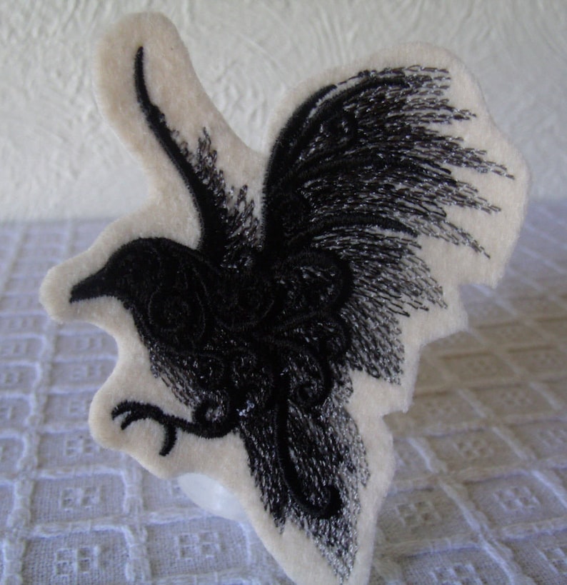May include: Black embroidered crow with detailed wings on a white felt background.