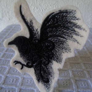 Peut inclure: Un oiseau noir brodé avec des détails complexes sur un fond de feutre blanc.