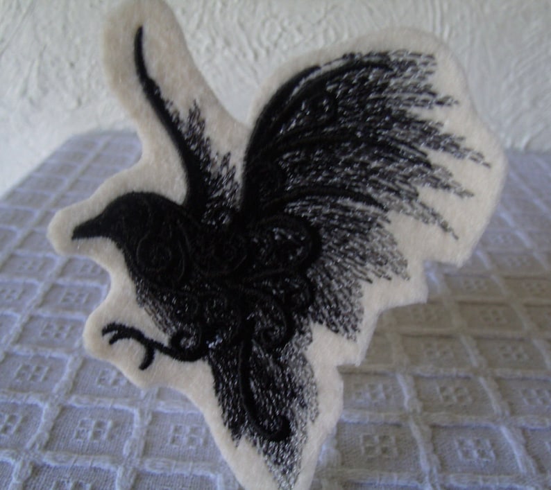 May include: A black embroidered bird with intricate details on a white felt background.
