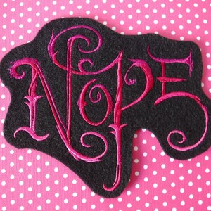 May include: A black felt patch with pink embroidery that reads "Nope" in a stylized font.