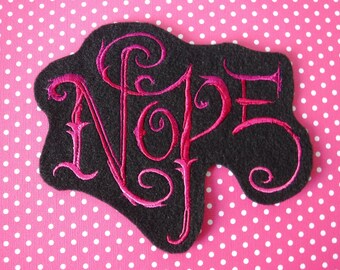 Peut inclure: Un patch en feutre noir avec une broderie rose qui dit "Nope" dans une police stylisée.