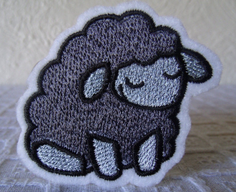 May include: A black embroidered sheep with a white outline and a white background. The sheep has a closed eye expression.